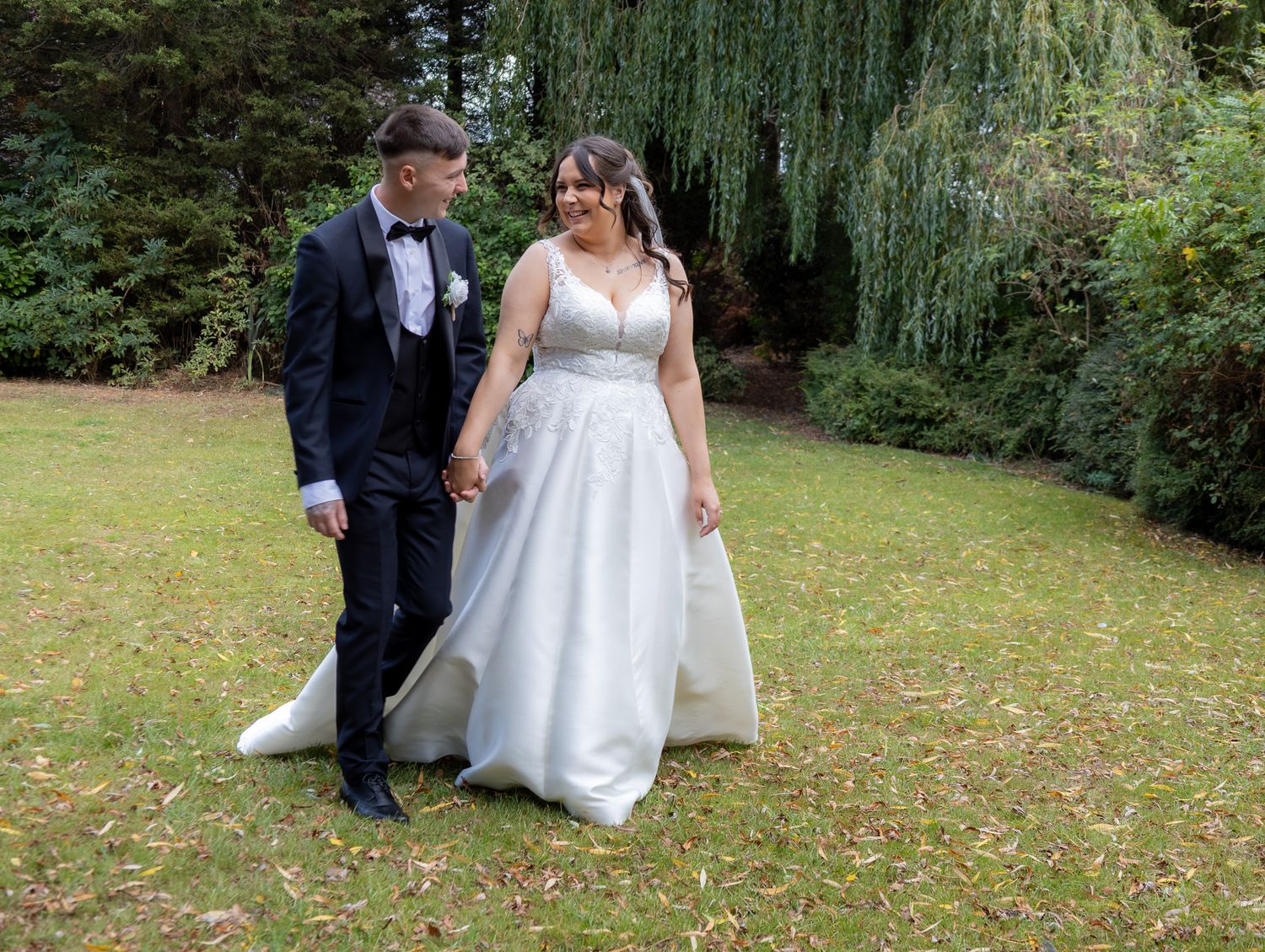 A couple in wedding attire walks together on a garden path surrounded by lush greenery and willow trees.