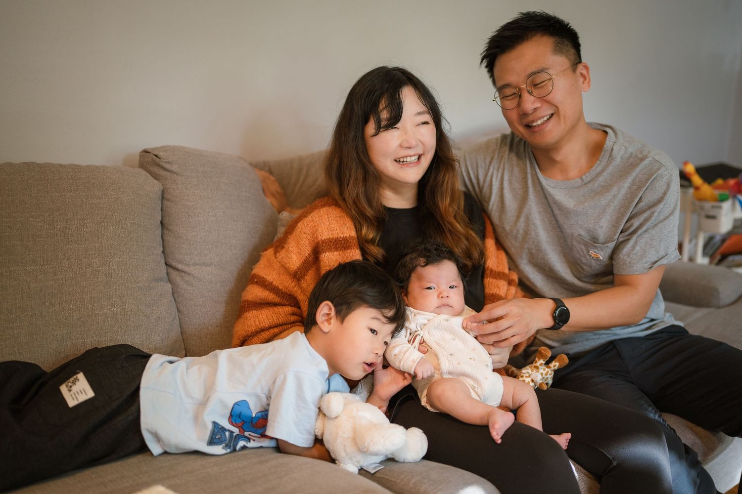 A family relaxes together on a grey couch, with the young child holding a stuffed toy and a baby in their midst.