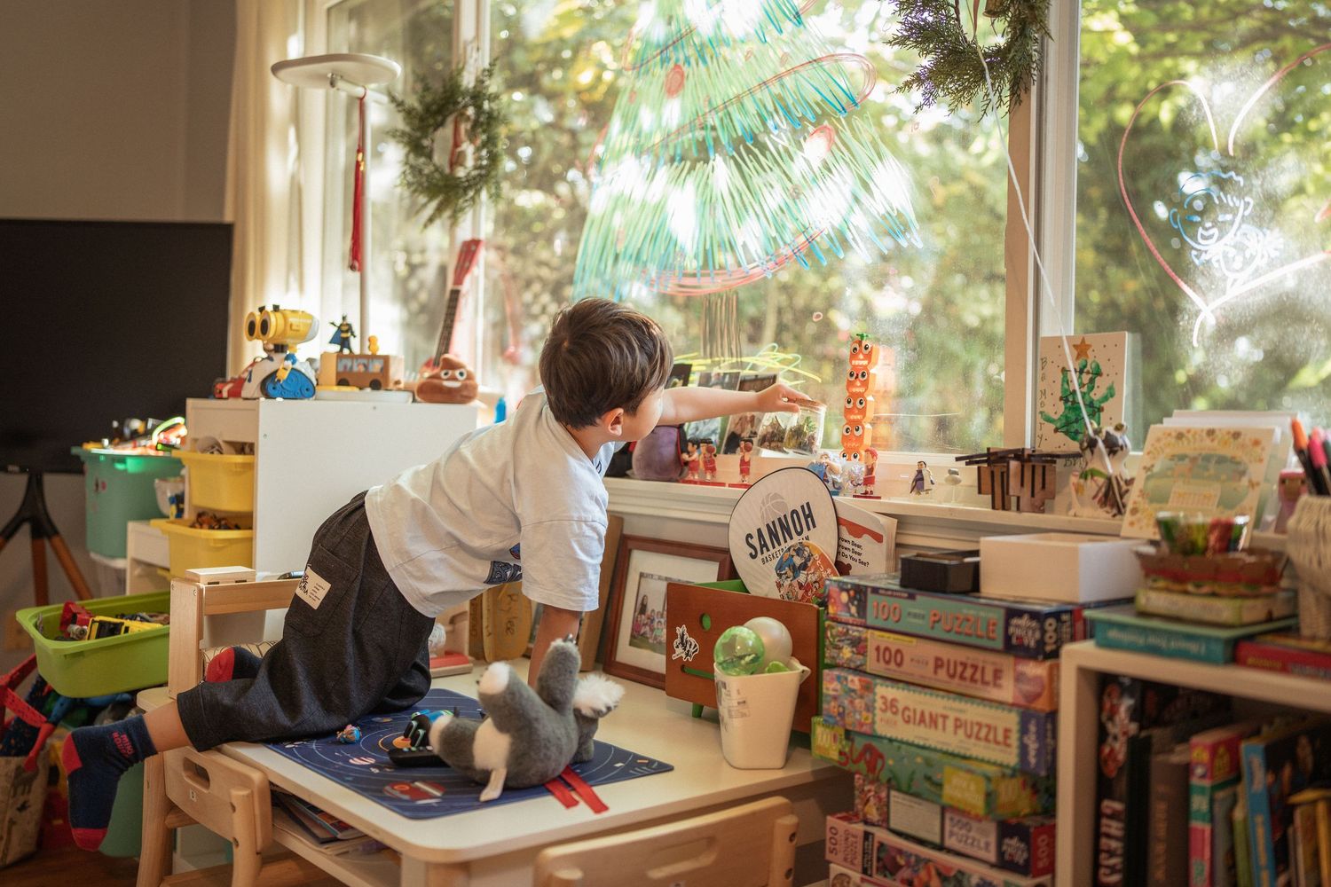 Sunlit living room features wooden furniture and toys scattered around large windows with natural greenery outside.