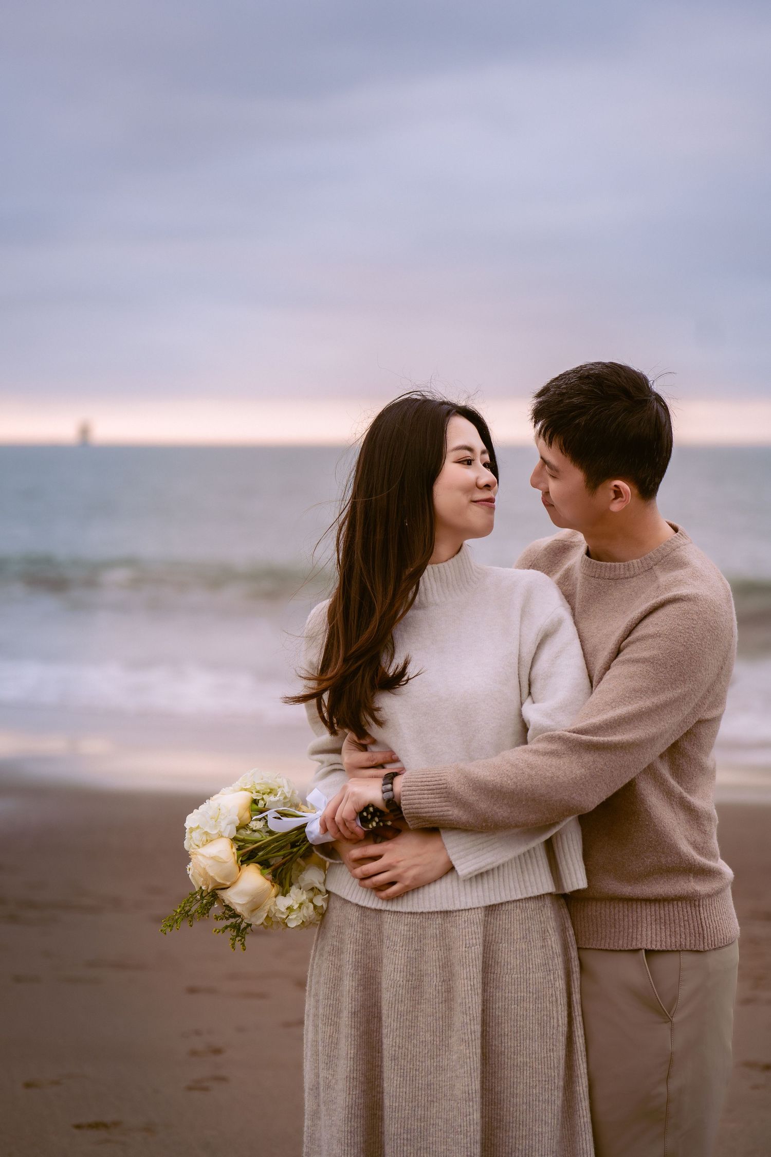 Couple in cozy beige sweaters sharing intimate moments on a misty beach at sunset.