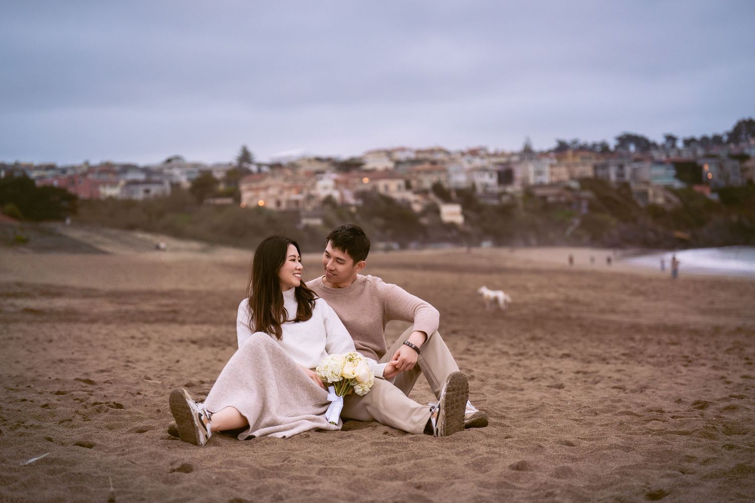 Romantic beach photoshoot with couple lounging on sand at sunset with bouquet of white flowers.