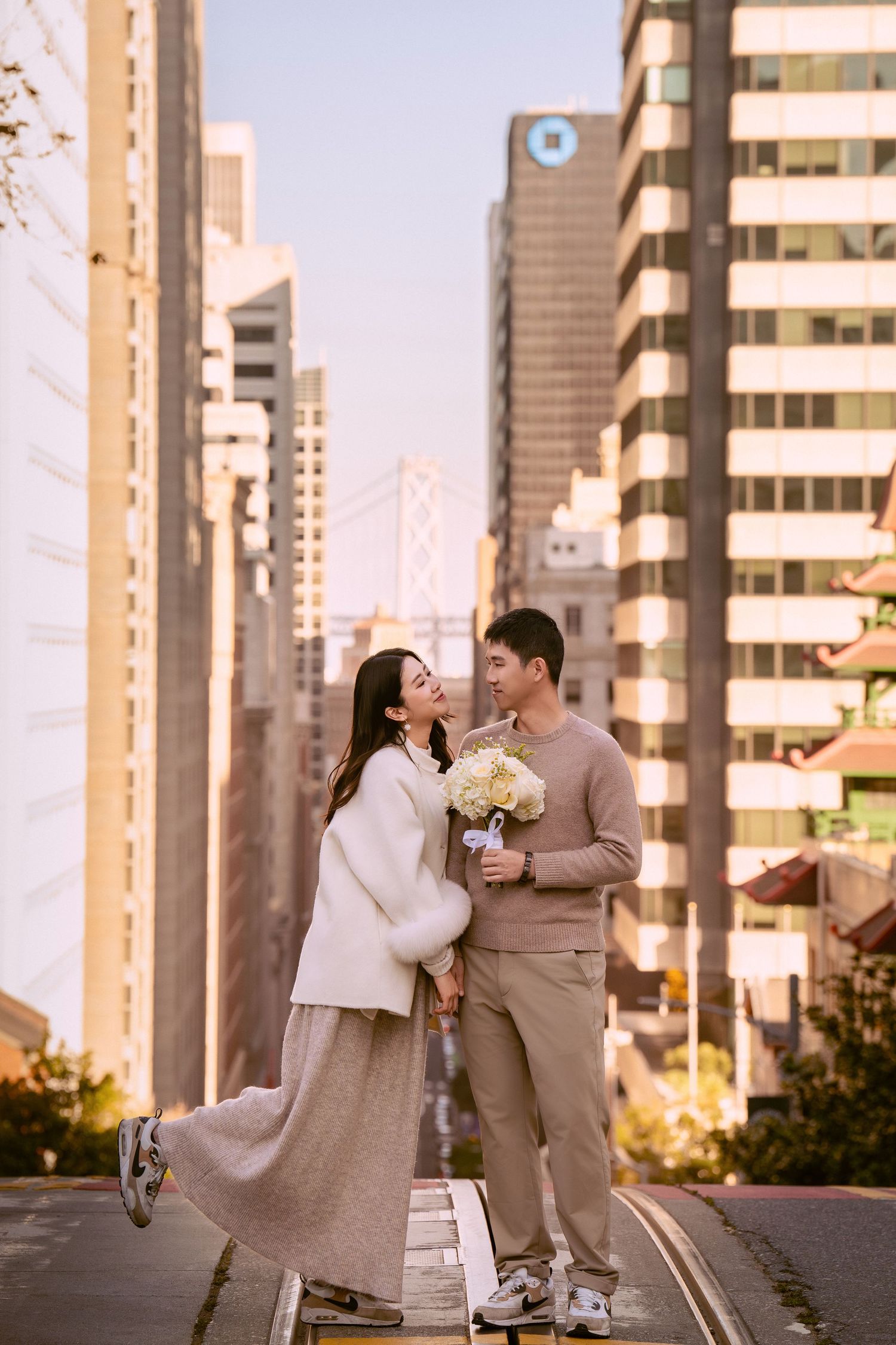 Couple in coordinated beige and white outfits share romantic moments against an urban backdrop at sunset.