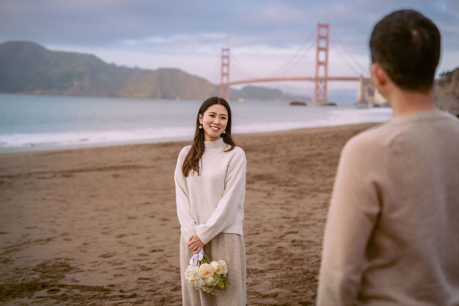 A couple shares a romantic moment on Baker Beach with the iconic Golden Gate Bridge in the background at sunset.