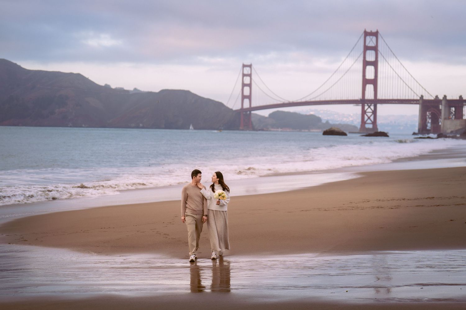 A couple walks along Baker Beach with the iconic Golden Gate Bridge visible in the foggy San Francisco background.