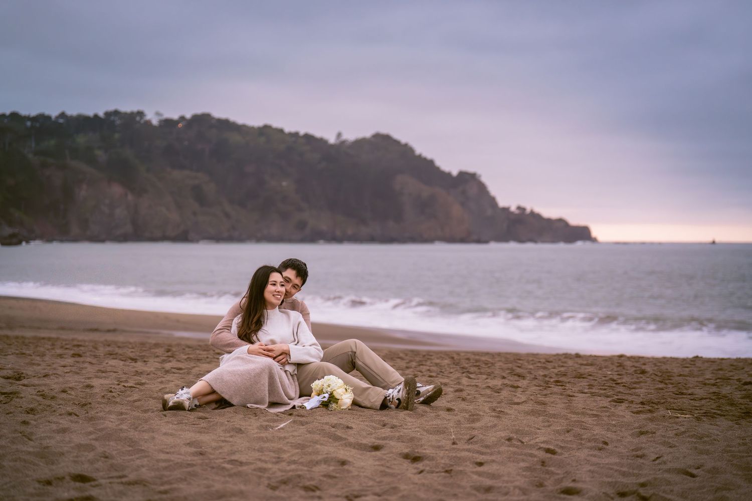 Romantic couple relaxing on a sandy beach at dusk with ocean waves and cliffs in the distance.