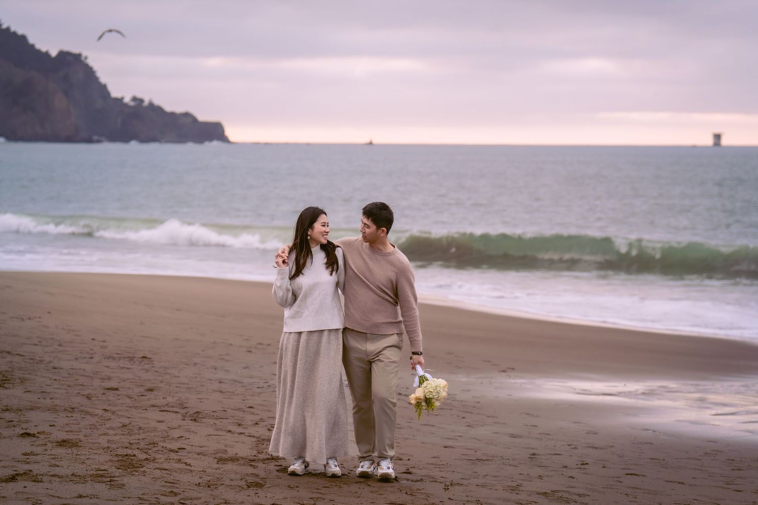 Romantic beach scene near the Golden Gate Bridge with figures walking along the sandy shoreline at sunset.