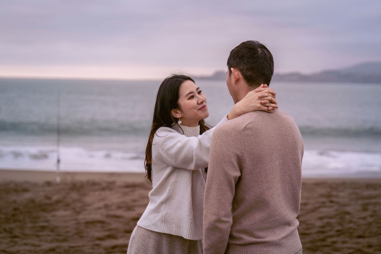 A romantic couple in neutral-toned outfits embraces on a misty beach at sunset.