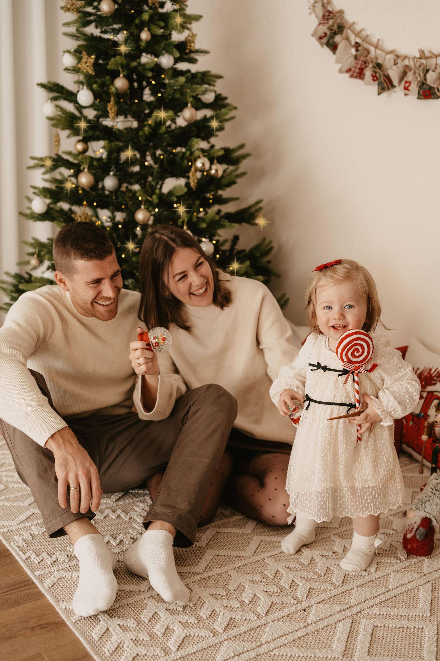 Family sits together on floor by Christmas tree sharing holiday moments in cozy neutral-toned setting.