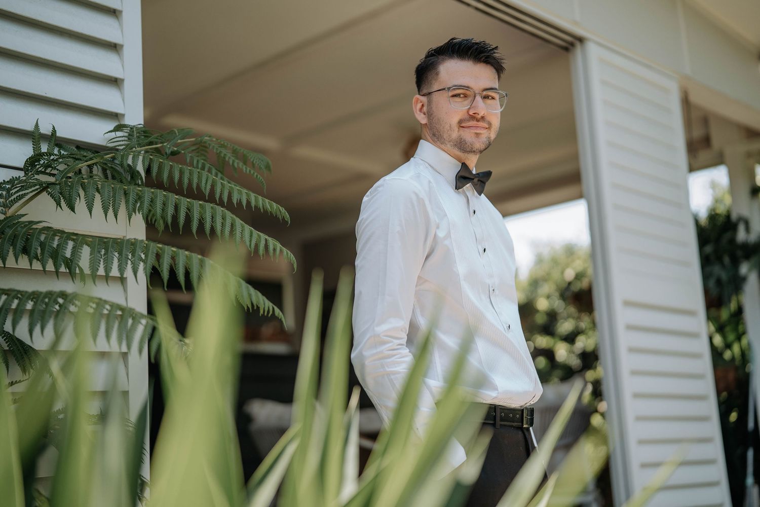 A person in a white dress shirt stands in a covered porch area surrounded by lush tropical plants and greenery.