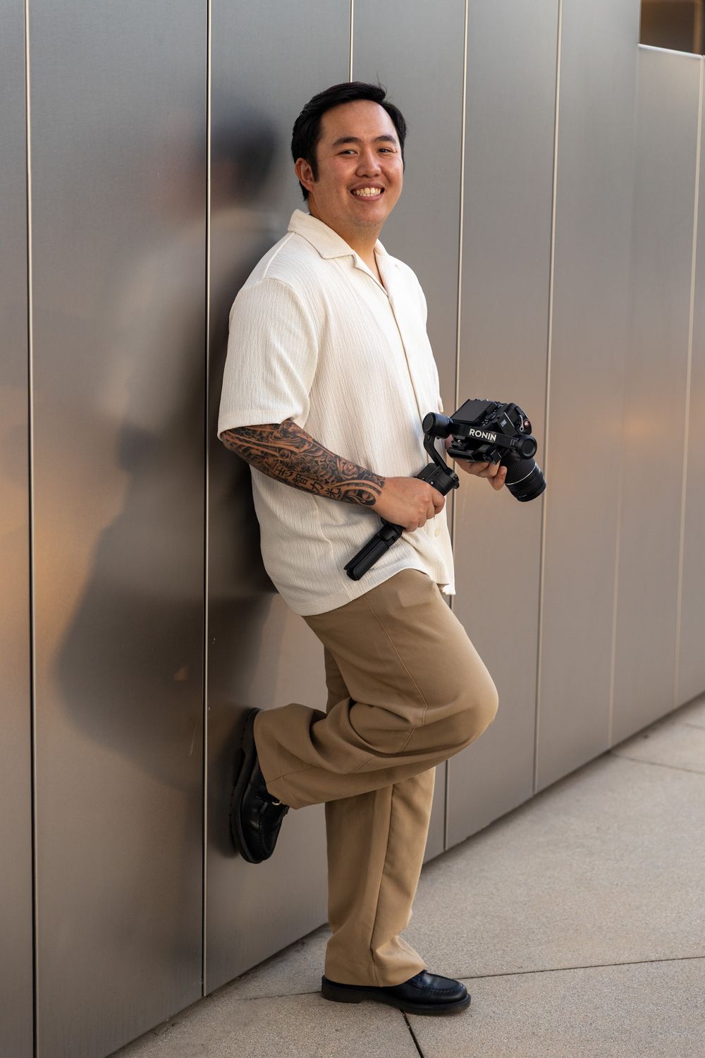 Professional photographer in casual attire holding a camera against a gray wall.
