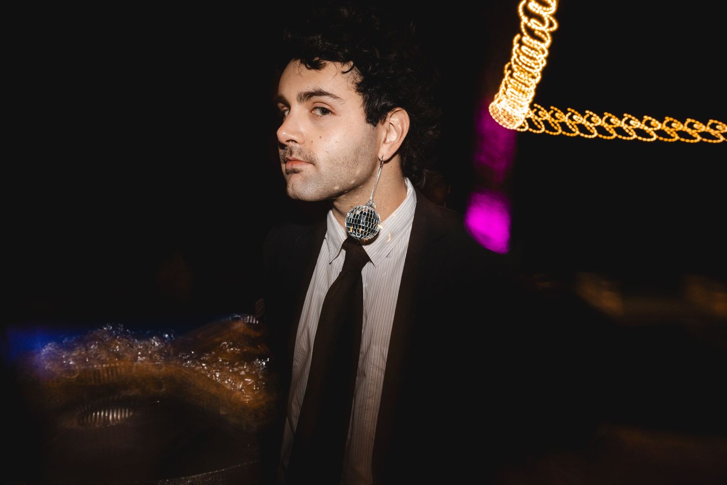 A person in a dark suit stands on stage against a black background with glowing lights in the distance.