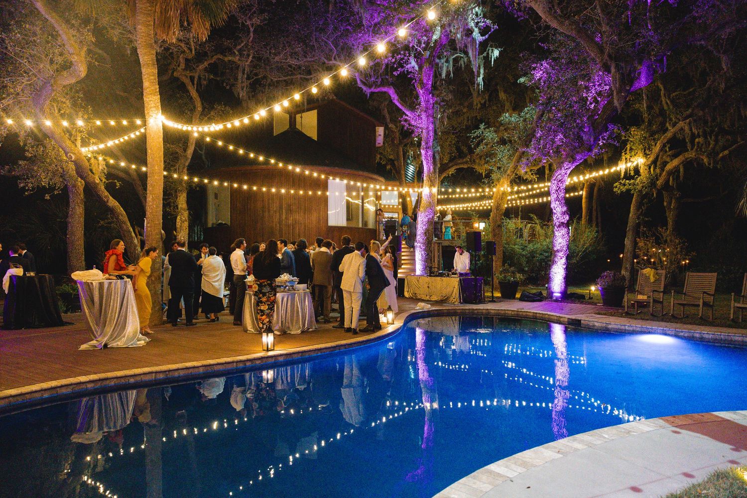String lights illuminate an outdoor pool area at night with purple uplighting creating a festive atmosphere.