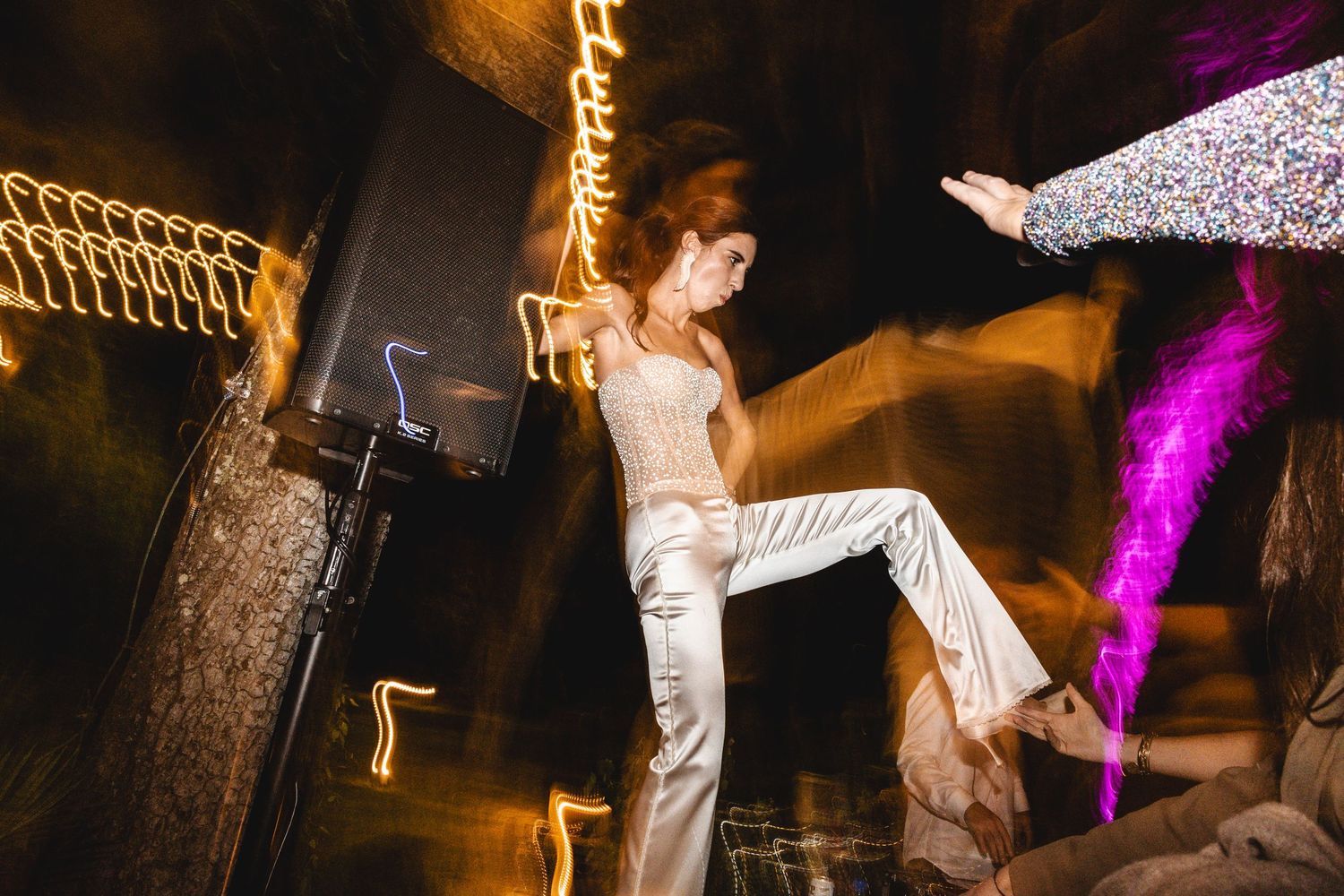 Wedding guests dance and celebrate under warm lighting and decorative string lights at an evening reception.