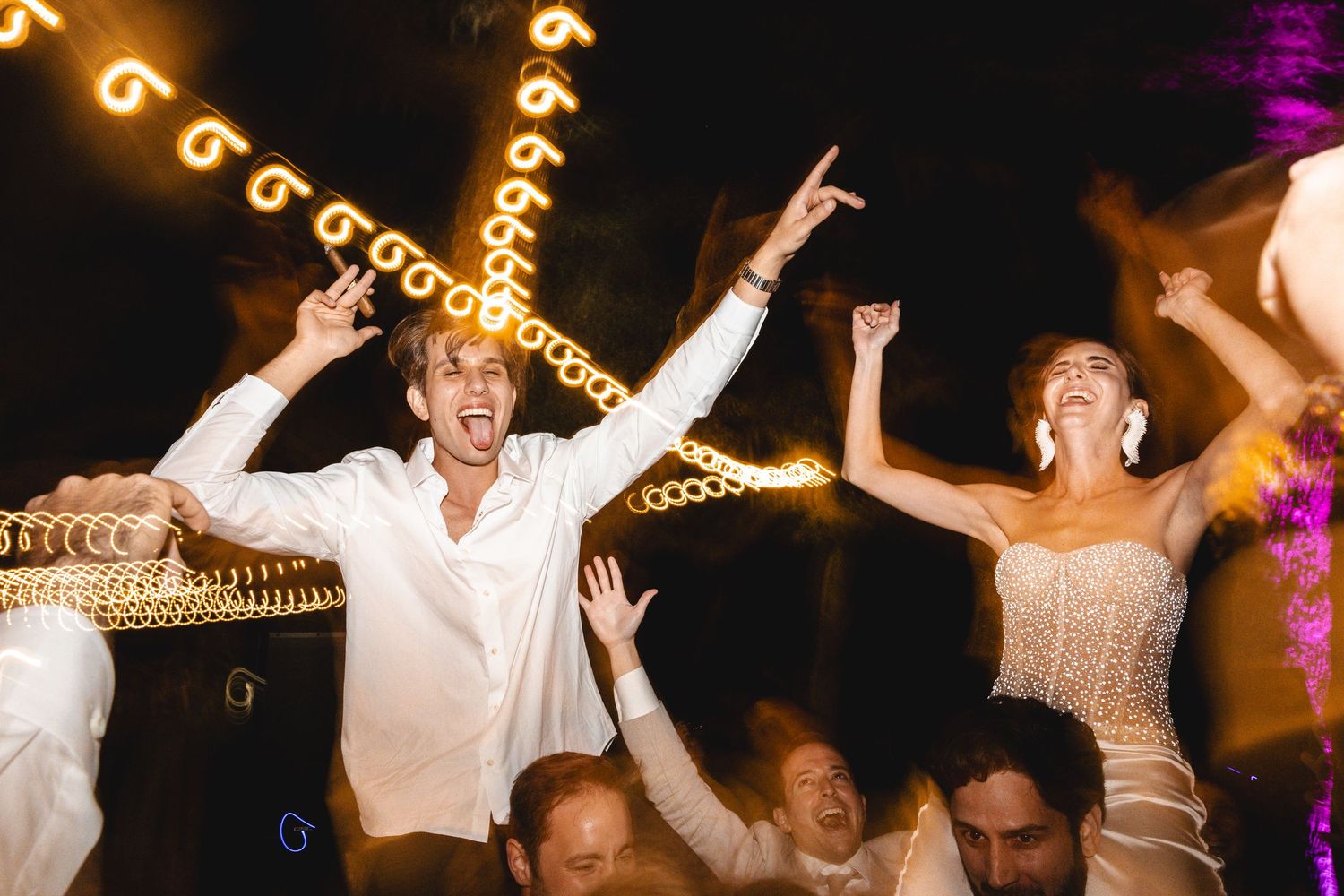 People dancing and celebrating at a wedding reception with string lights glowing overhead in the evening.