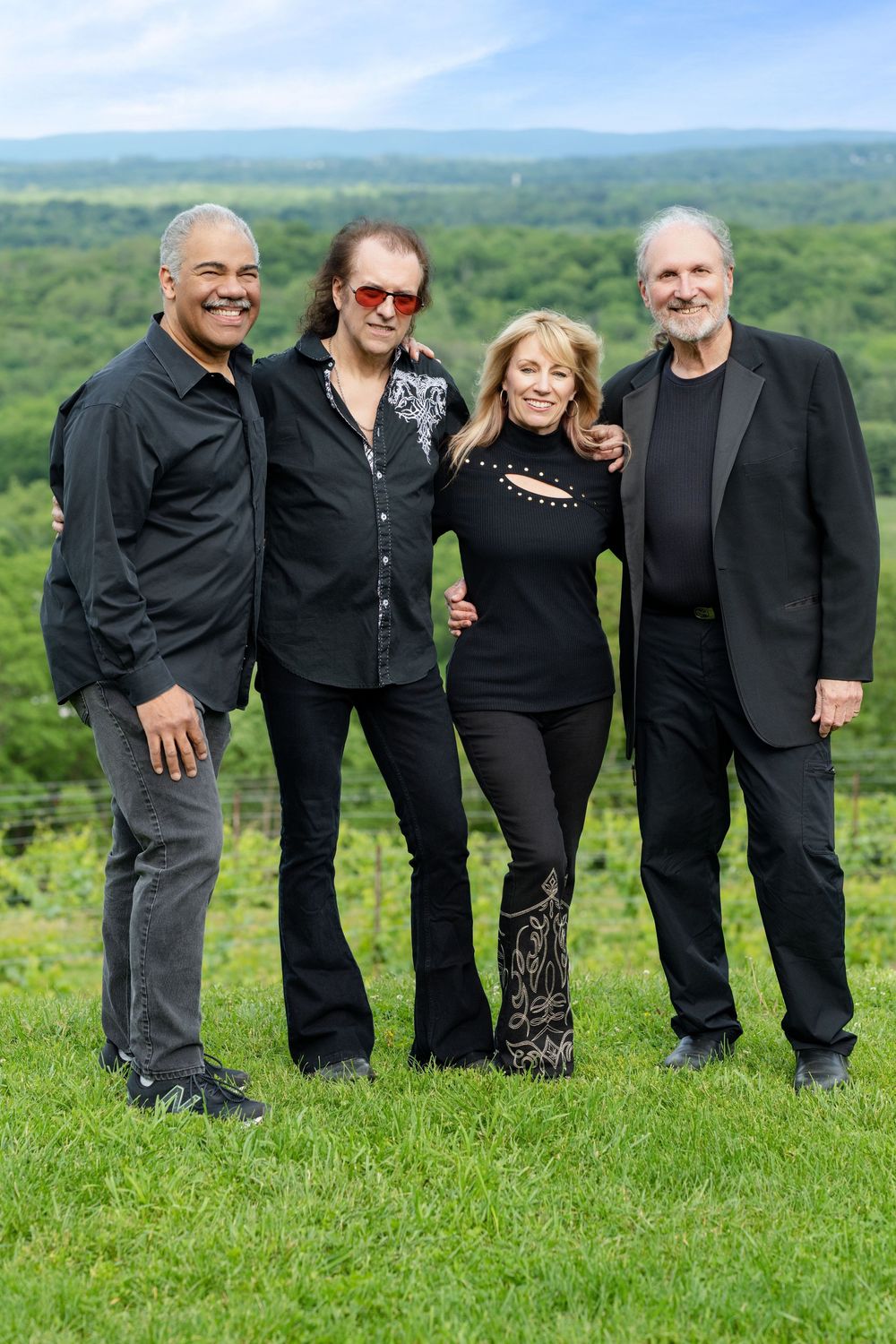 Four people in black attire standing together on hilltop with scenic forest view.