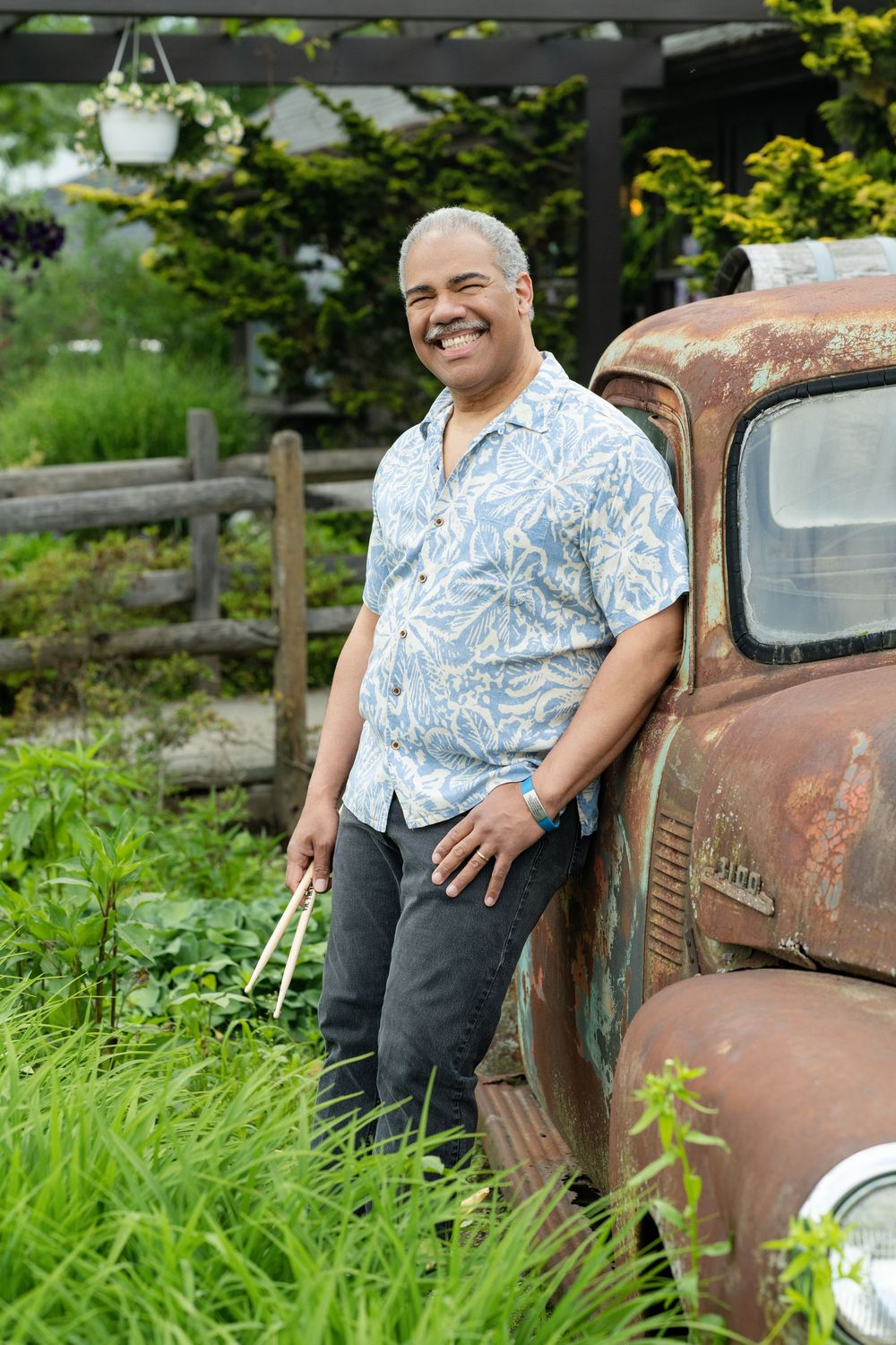 Person in Hawaiian shirt poses casually against rustic vintage truck in lush garden setting.