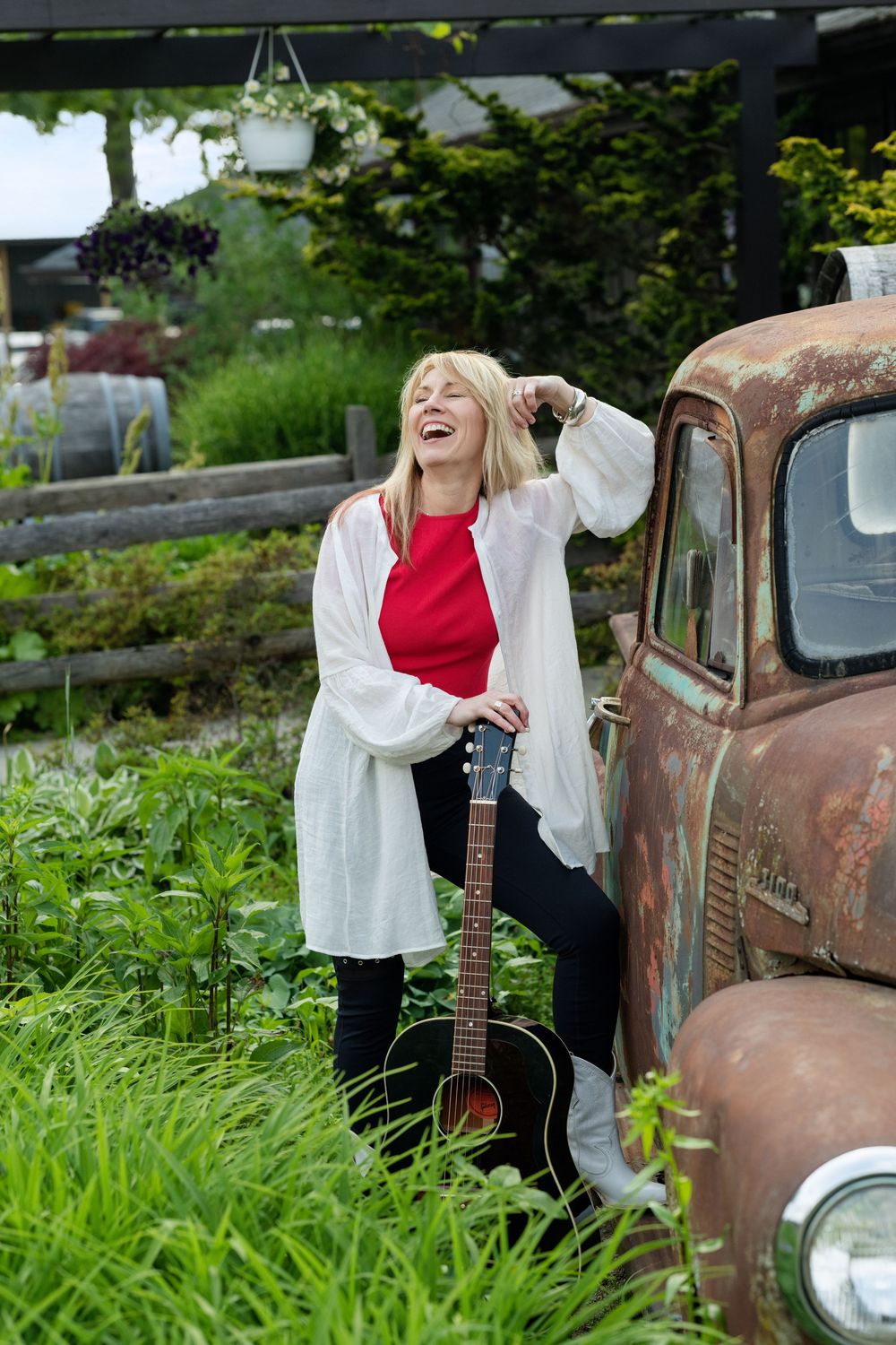 Musician in red top and white cardigan laughs while posing with acoustic guitar next to rustic vintage truck.