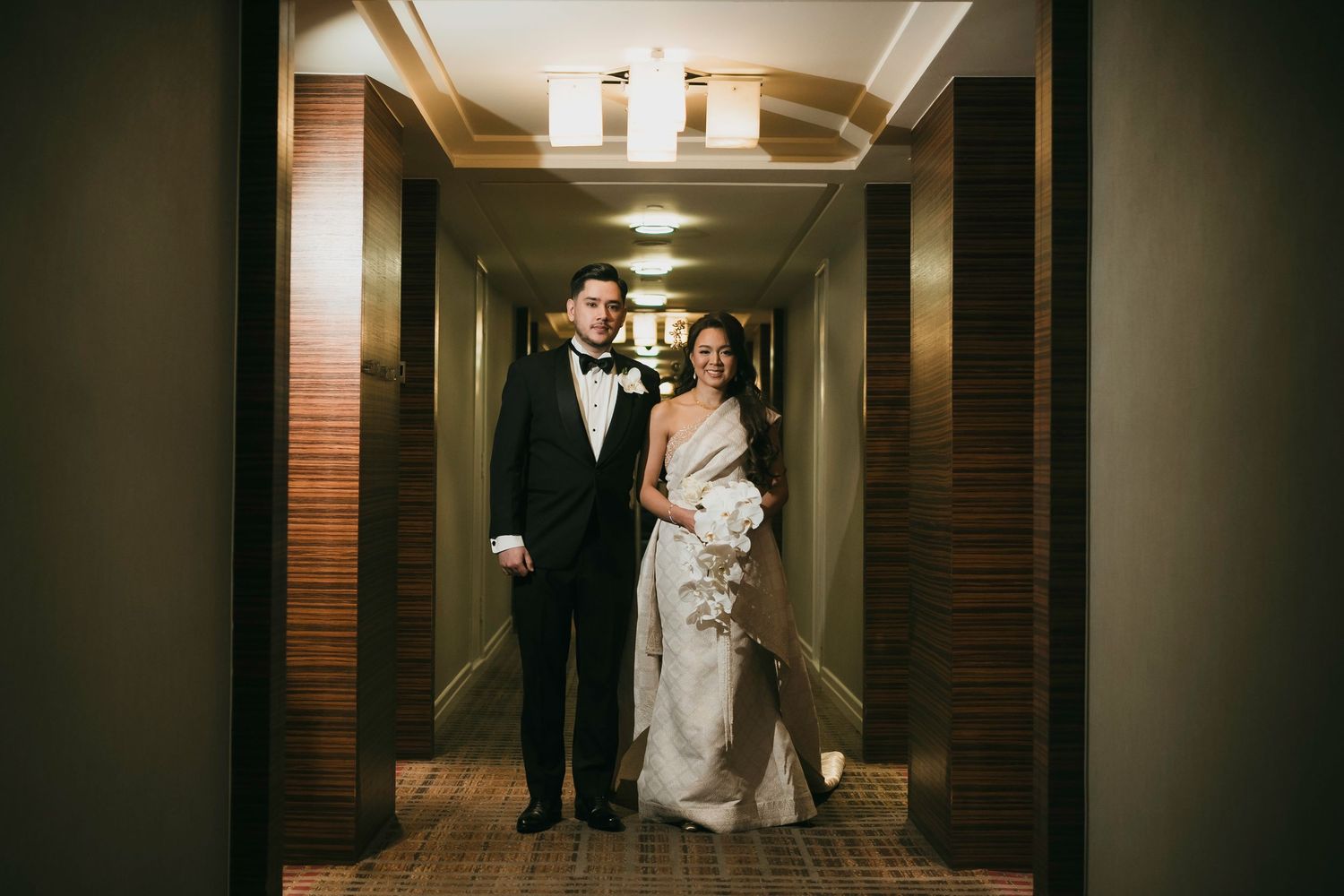Bride in elegant sari and groom in tuxedo pose together in a warmly lit hotel corridor on their wedding day.