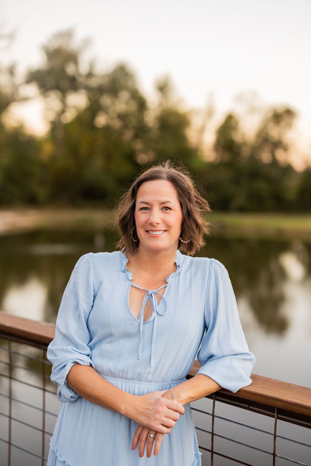 A person in a light blue dress smiles while standing on a deck overlooking a pond at sunset.