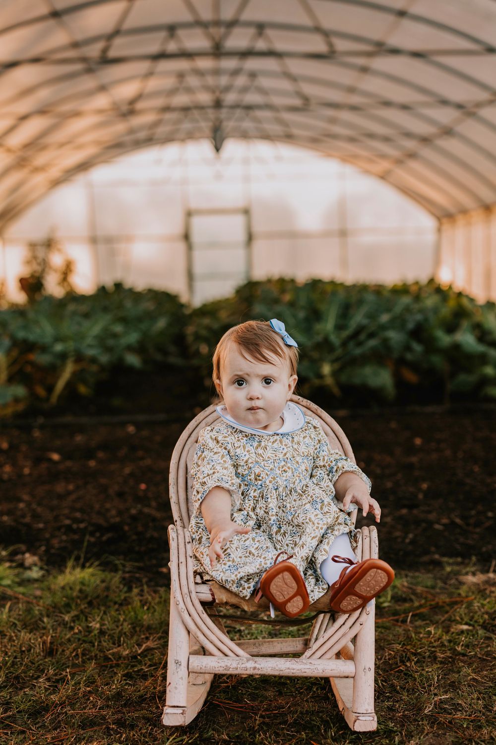 Small child sits in a pink rocking chair wearing a floral dress inside a greenhouse.