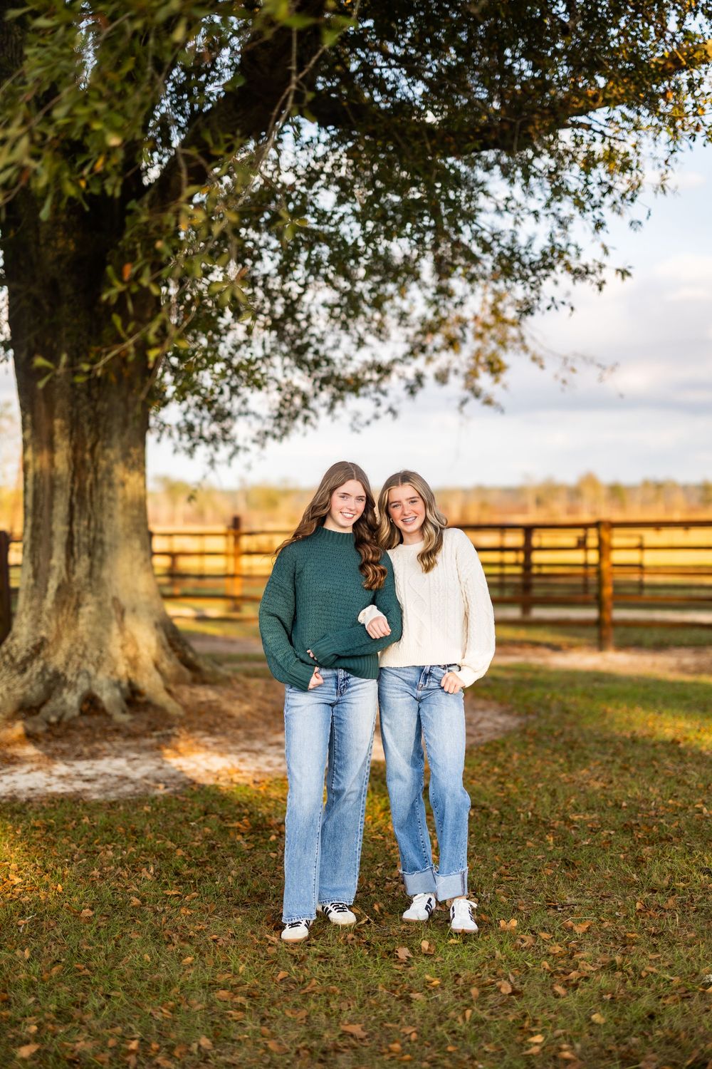 Two friends pose together under large tree on autumn farm.
