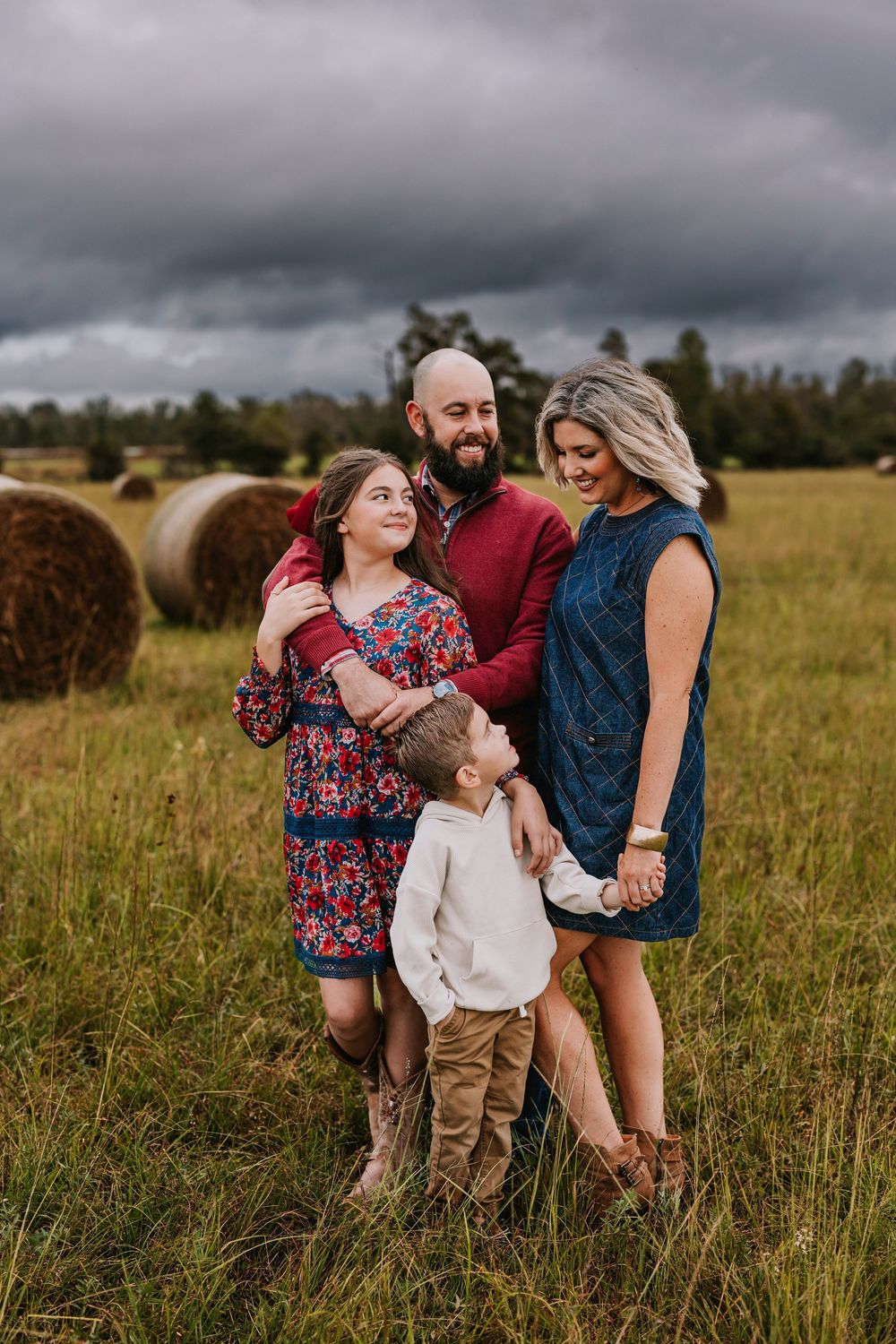 A family of four poses together in a field with hay bales and stormy skies in the background.