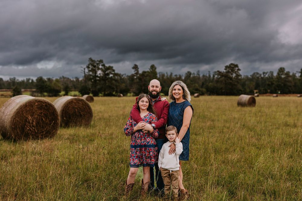 Family portrait in a rustic field with round hay bales and dramatic storm clouds overhead.