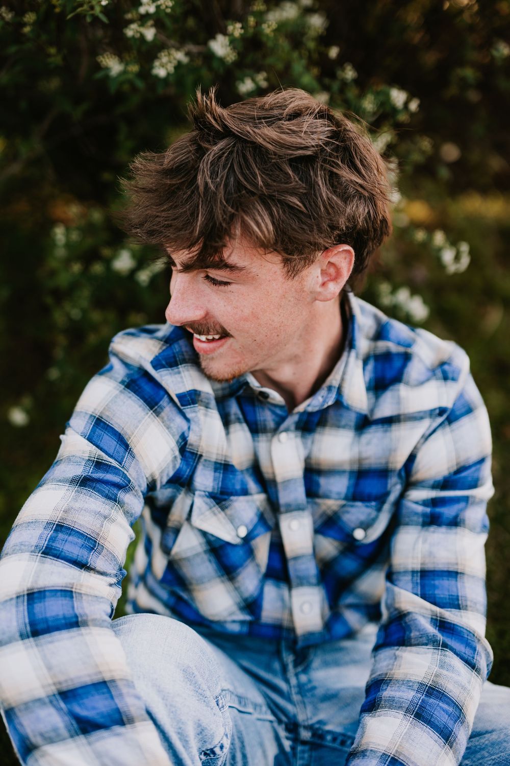 A candid portrait of someone in a blue plaid shirt smiling against a blurred autumn background.