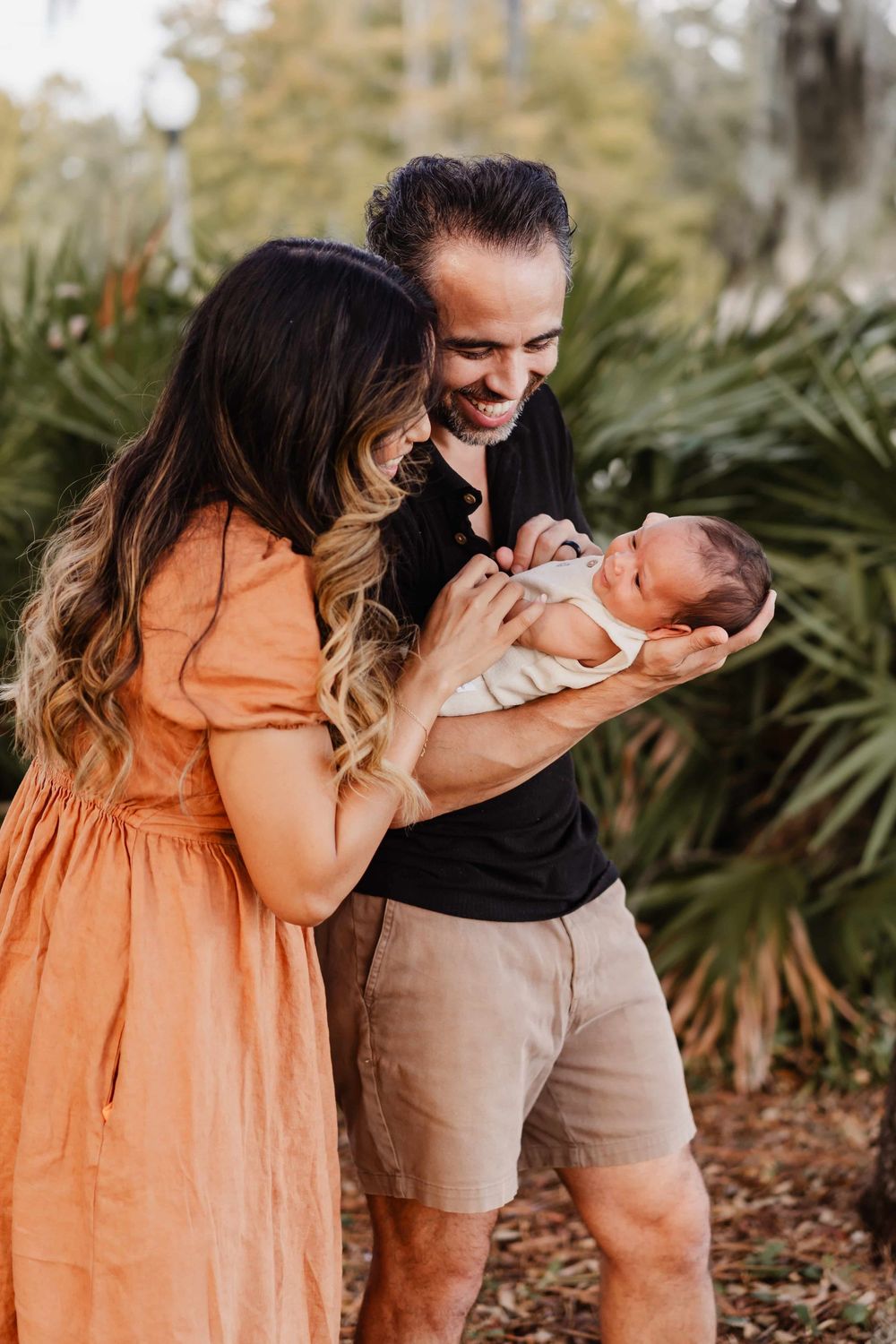 A couple in a garden setting share a joyful moment with their newborn baby against a backdrop of tropical plants.