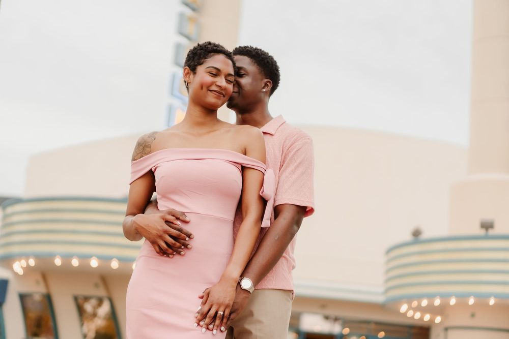 A romantic Black couple in pink outfits share a tender moment outdoors, the man kissing the womans cheek gently.