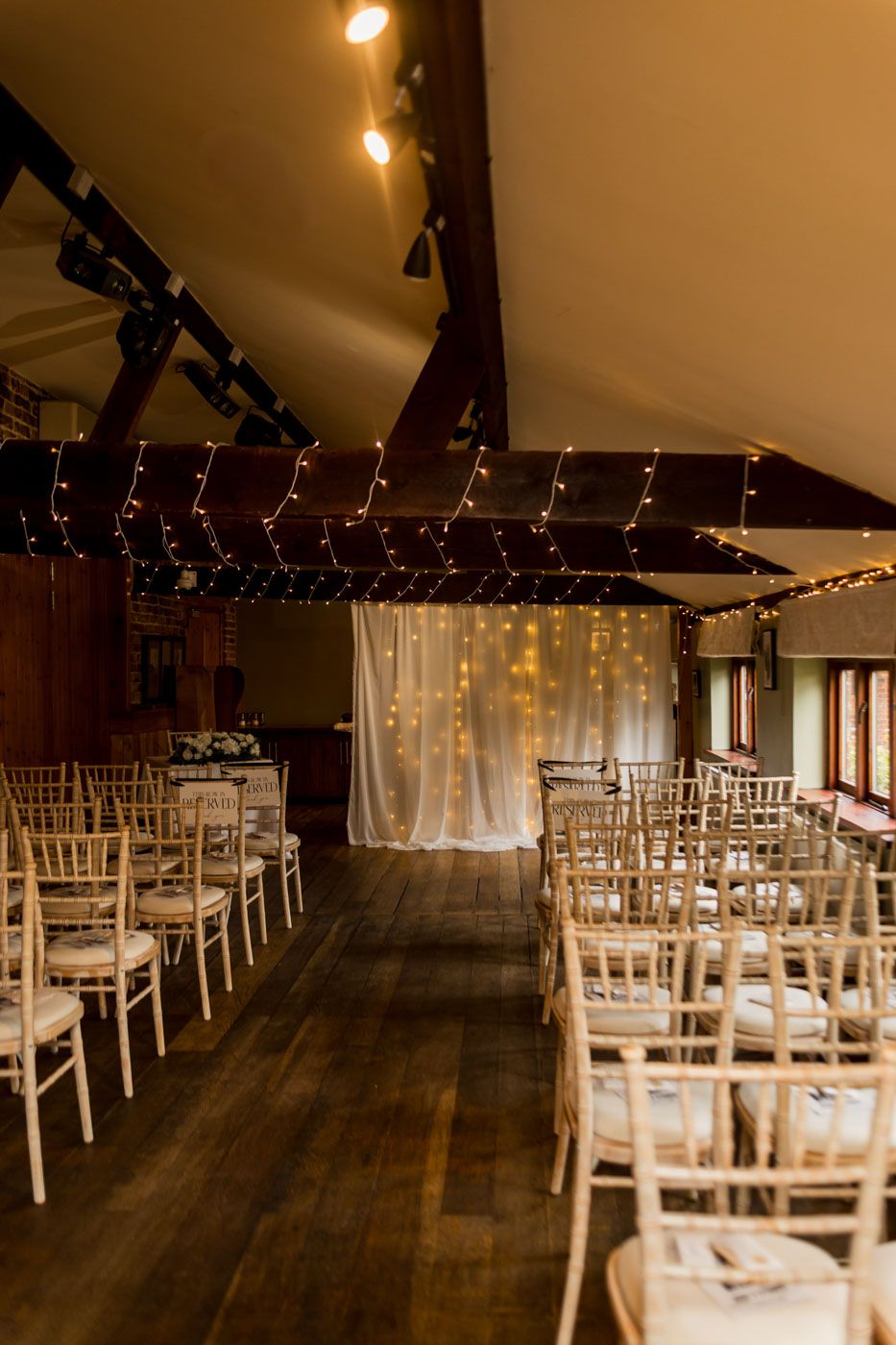 Rustic indoor wedding ceremony space with white chiavari chairs and fairy light decorations.