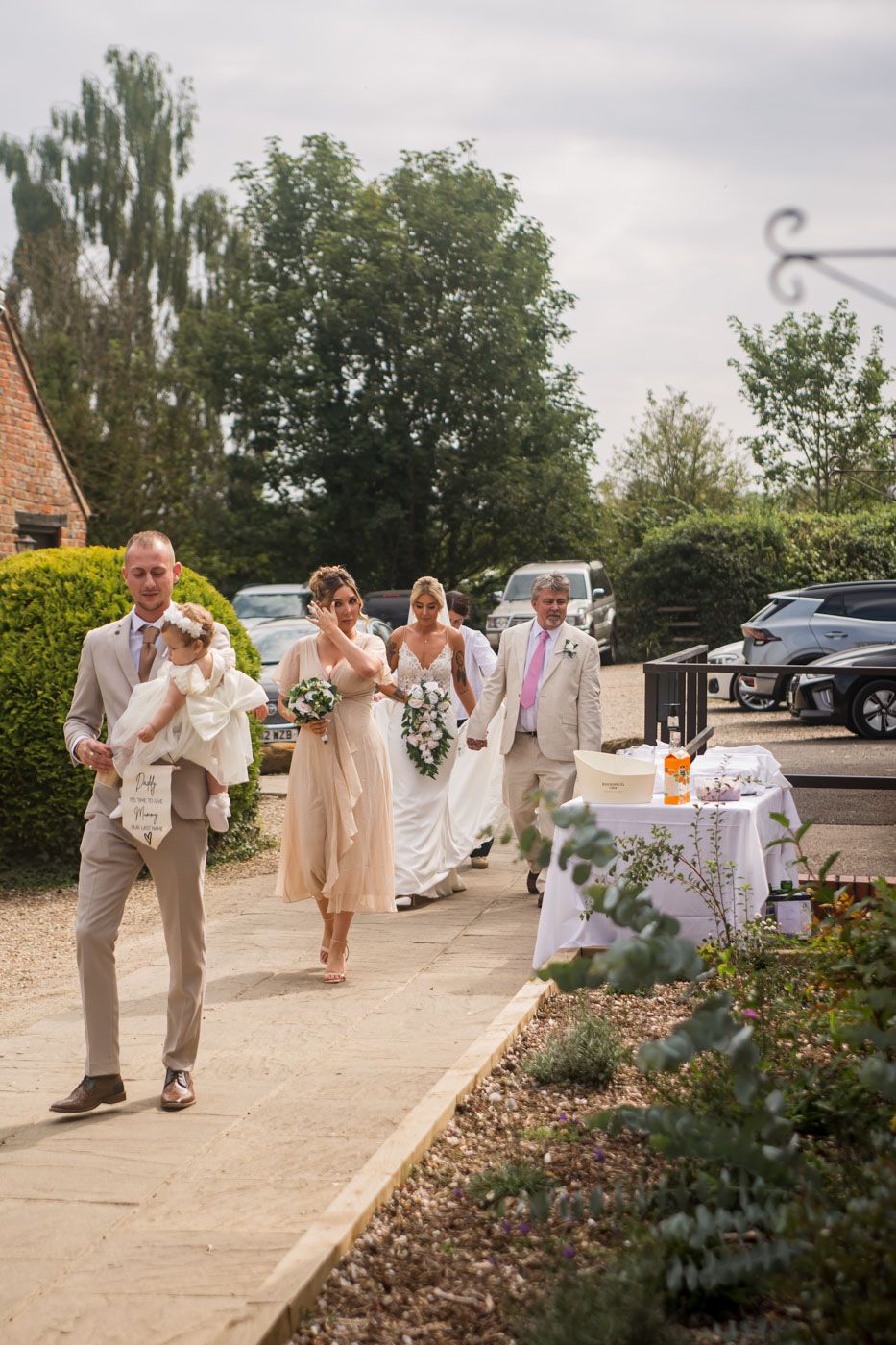 Wedding party members walking and celebrating on outdoor pathway near parked cars.
