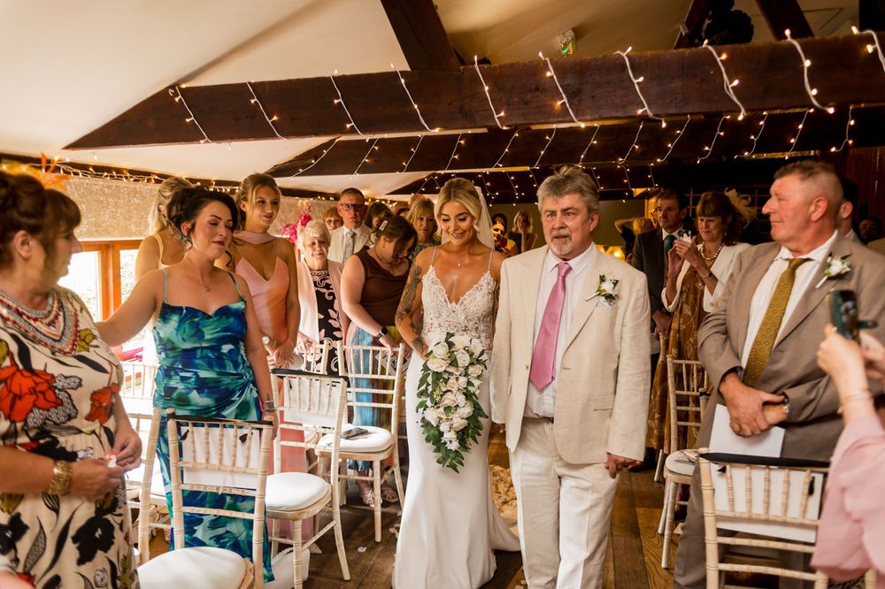 Wedding attendees seated in white chairs under string lights during an indoor ceremony.