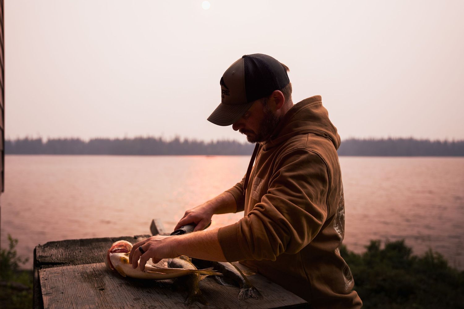 A person in a brown hoodie and cap prepares food at an outdoor counter during sunset.
