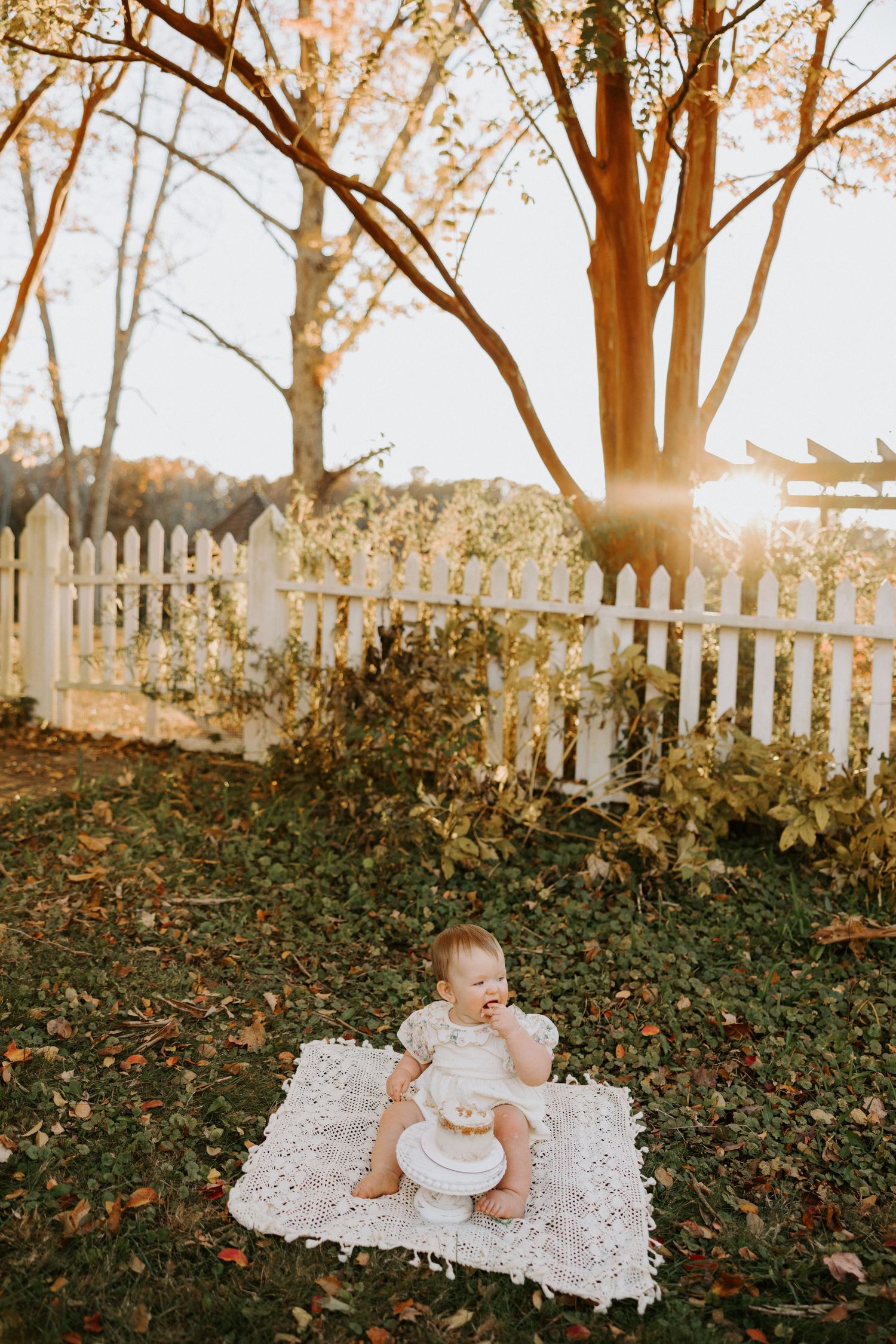White lace blanket laid on autumn leaves with sunlight filtering through trees and white picket fence in background.