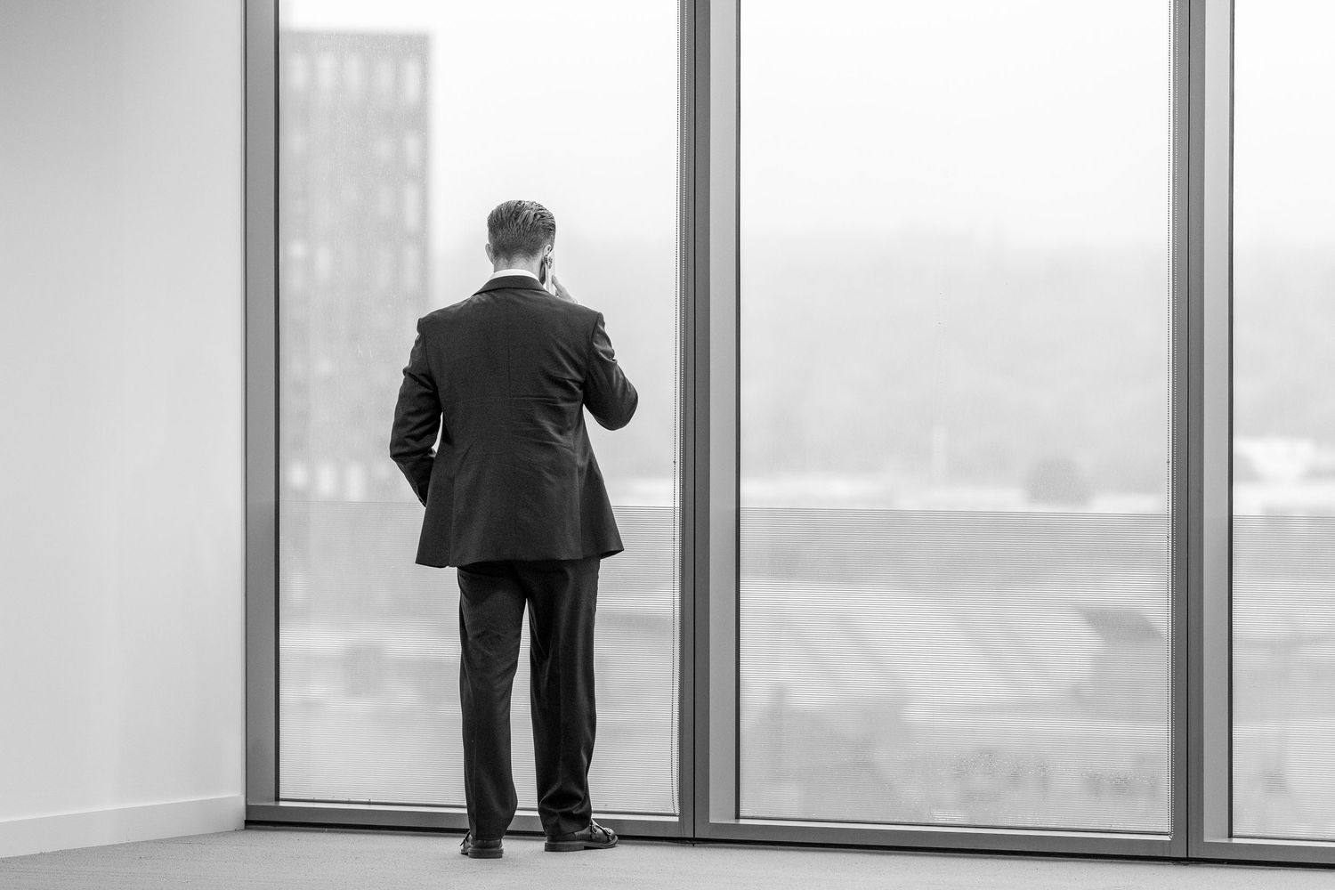 A businessman in a dark suit stands at a large window overlooking a city skyline in black and white.