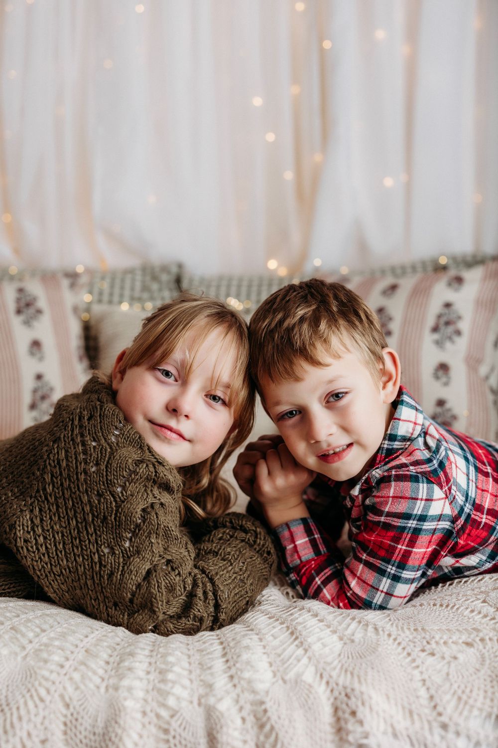 A sequence of five cozy holiday photos showing two young siblings cuddling together on a white blanket with twinkling lights.