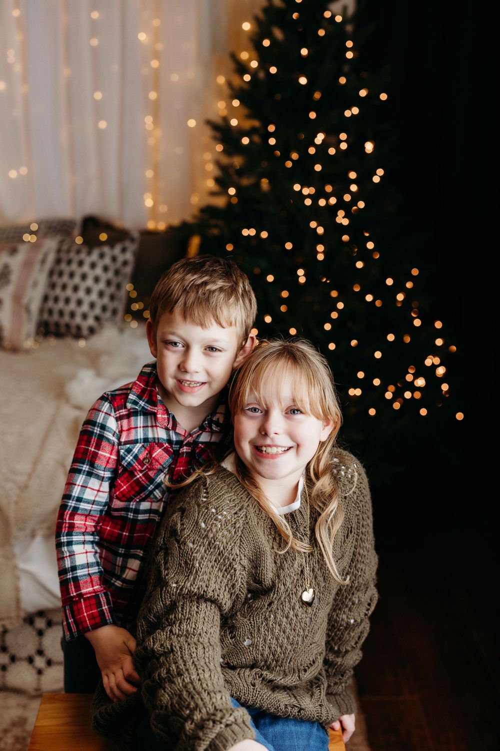 Two children pose playfully in cozy winter outfits with twinkling Christmas lights and decorations in the background.