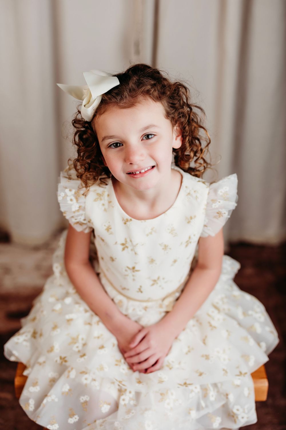 A child in a white and gold floral dress with ruffled sleeves sits and smiles while posing for multiple photos.