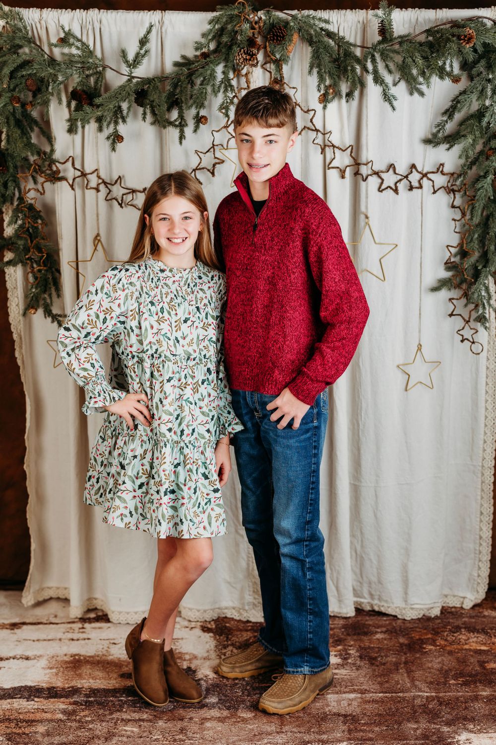 Two children pose in holiday attire with evergreen garland and gold star decorations against a white backdrop.