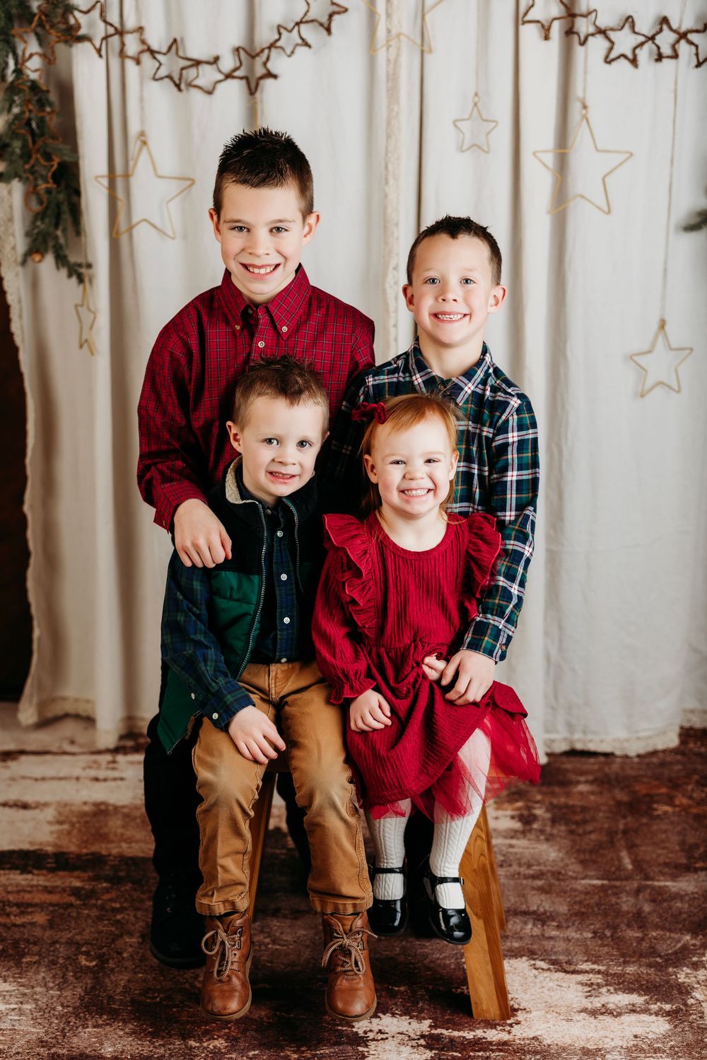 Three siblings pose together in coordinated holiday outfits against a rustic backdrop with hanging star decorations.