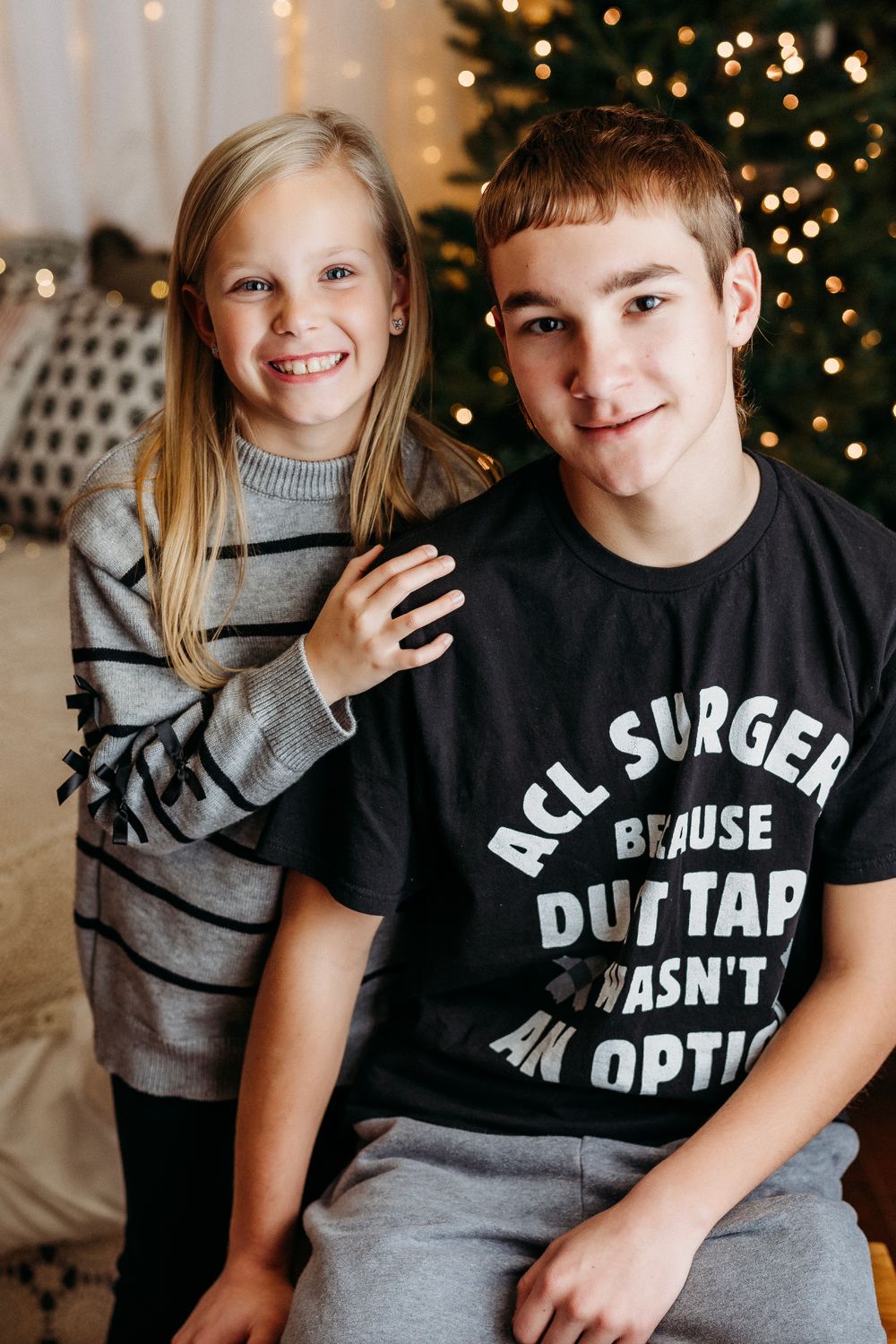 Two children sit close together smiling in front of a Christmas tree with twinkling lights in the background.