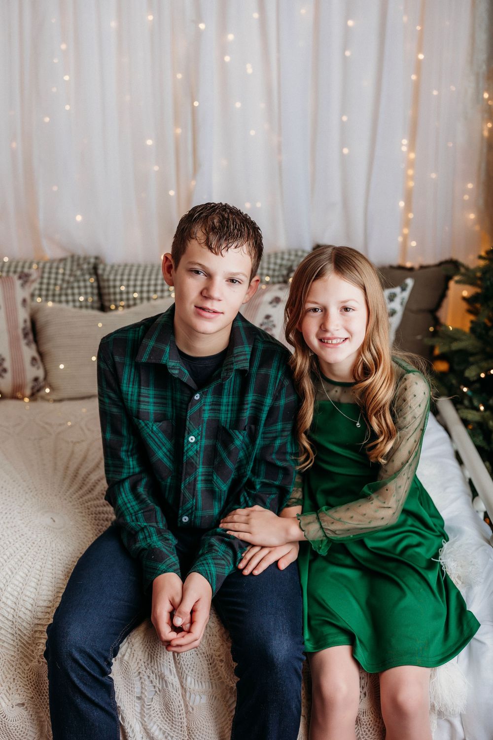 A festive holiday photoshoot with siblings wearing coordinated green and plaid outfits against a twinkle light backdrop.