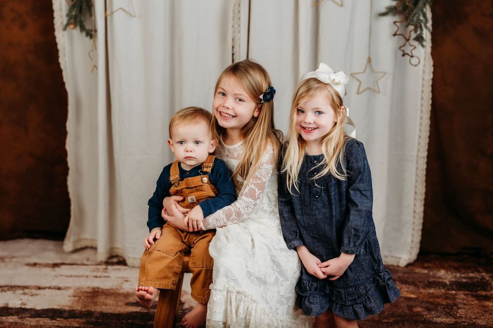 A group of children pose together in coordinating outfits against a rustic backdrop with white curtains.