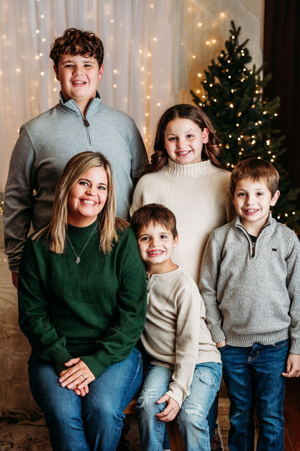 A family poses together in coordinating sweaters in front of a Christmas tree and twinkle light backdrop.
