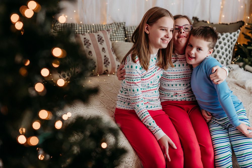 Family wearing matching holiday pajamas sits together on couch with Christmas tree lights glowing in background.