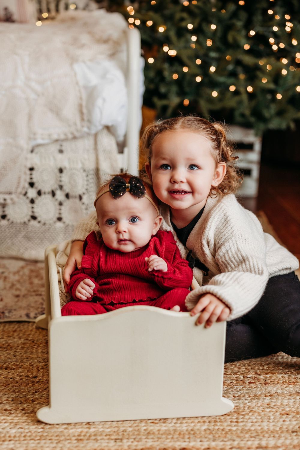 Two small children pose together by a Christmas tree with one wearing red and one in cream colored sweater.