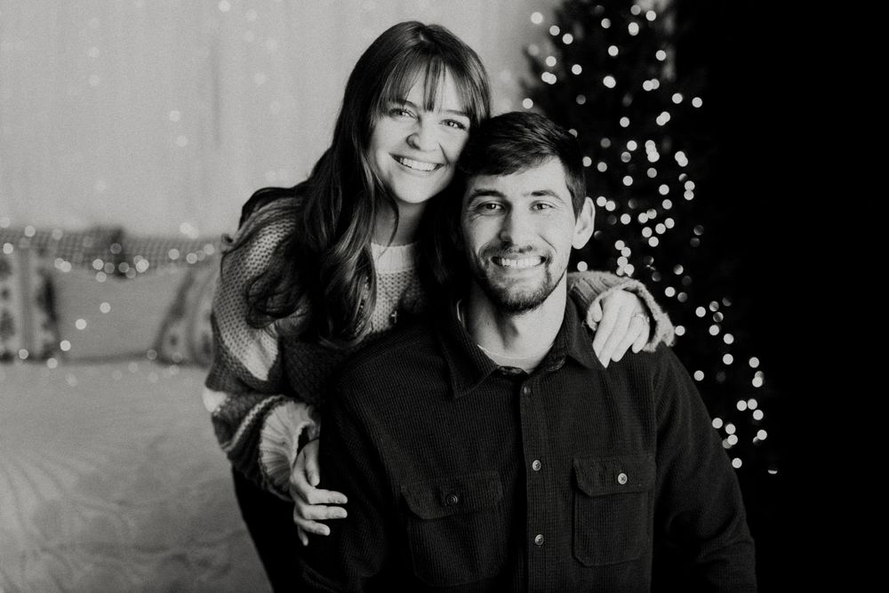 A couple smiles and embraces during a winter photoshoot with twinkling lights in the background.