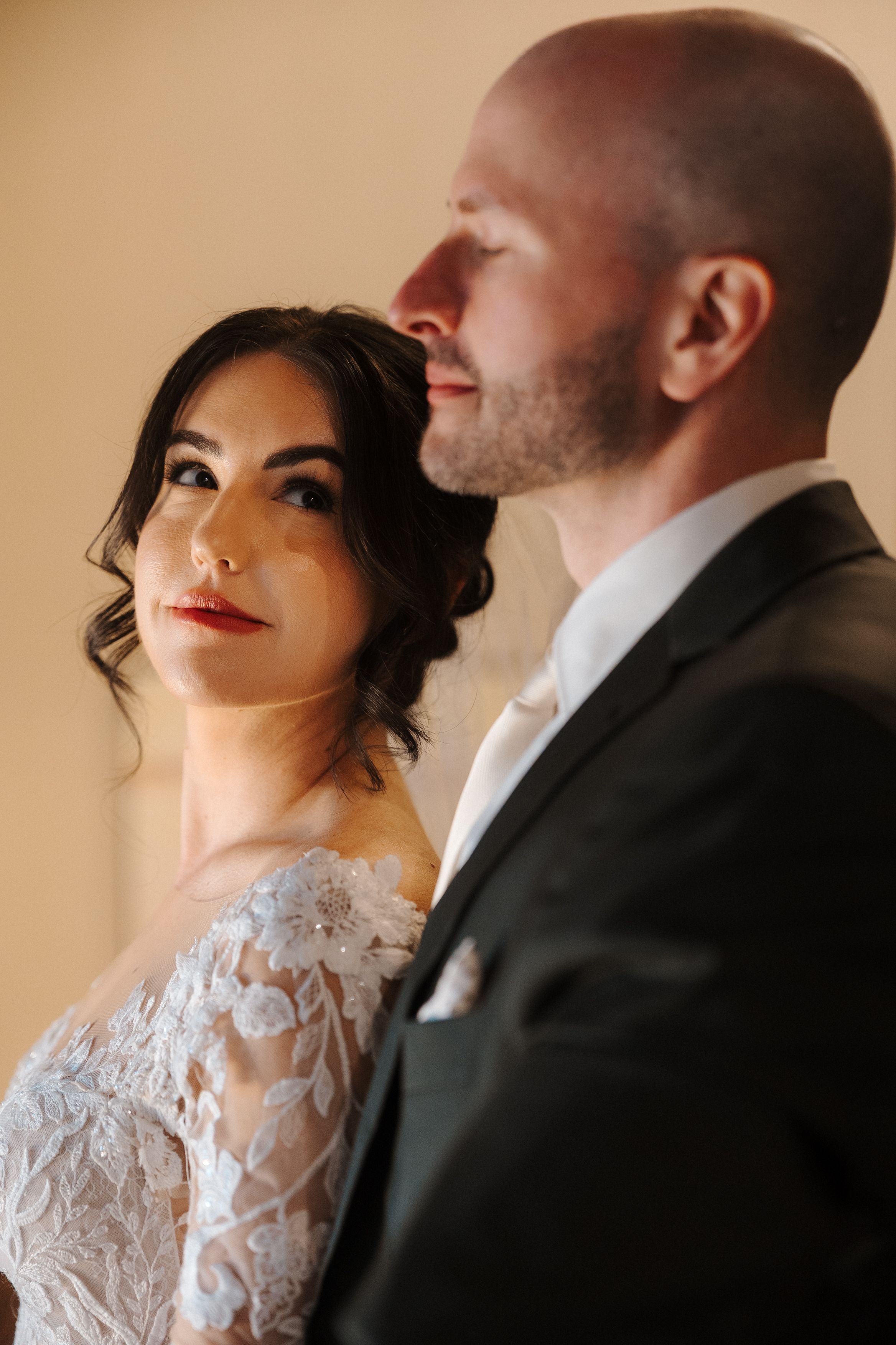 A romantic wedding couple in intimate poses with the bride wearing a delicate lace dress against a beige background.