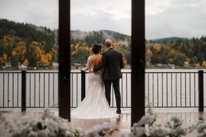 Wedding couple stands on a deck overlooking a scenic mountain lake with autumn foliage.